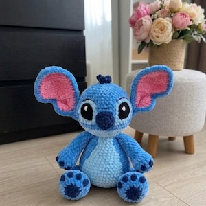 Puede incluir: Un peluche de ganchillo hecho a mano del personaje Stitch, con un cuerpo azul brillante, un parche en el pecho azul claro, orejas grandes con detalles interiores rosas, una nariz azul oscuro y ojos negros y blancos, sentado.