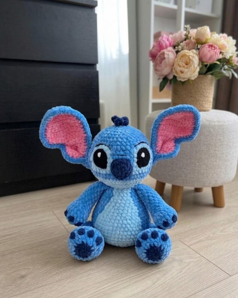 Puede incluir: Un peluche de ganchillo hecho a mano del personaje Stitch, con un cuerpo azul brillante, un parche en el pecho azul claro, orejas grandes con detalles interiores rosas, una nariz azul oscuro y ojos negros y blancos, sentado.