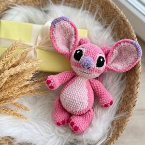 Puede incluir: Un peluche de ganchillo en rosa y rosa claro con orejas grandes de puntas moradas, nariz morada clara y ojos negros. La criatura rellena tiene un cuerpo redondo, extremidades cortas con almohadillas rosa oscuro y una peque&ntilde;a sonrisa.