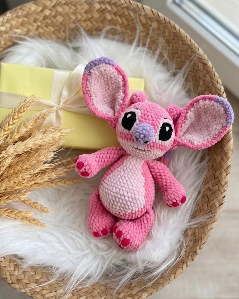 Puede incluir: Un peluche de ganchillo en rosa y rosa claro con orejas grandes de puntas moradas, nariz morada clara y ojos negros. La criatura rellena tiene un cuerpo redondo, extremidades cortas con almohadillas rosa oscuro y una peque&ntilde;a sonrisa.