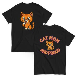 Może przedstawiać: Dwie czarne koszulki z pomarańczowymi grafikami kotów. Jedna koszulka przedstawia kota liżącego łapę, a druga napis "Cat Mom and Proud" z ilustracją kota. Koszulki są prawdopodobnie wykonane z bawełny.
