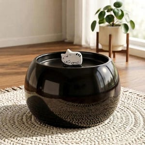 May include: A glossy black ceramic pet water fountain with a rounded, bowl-like shape. A small, white, hand-painted ceramic cat figurine with black stripes and a smiling expression sits on the centre of the lid, serving as a decorative topper.