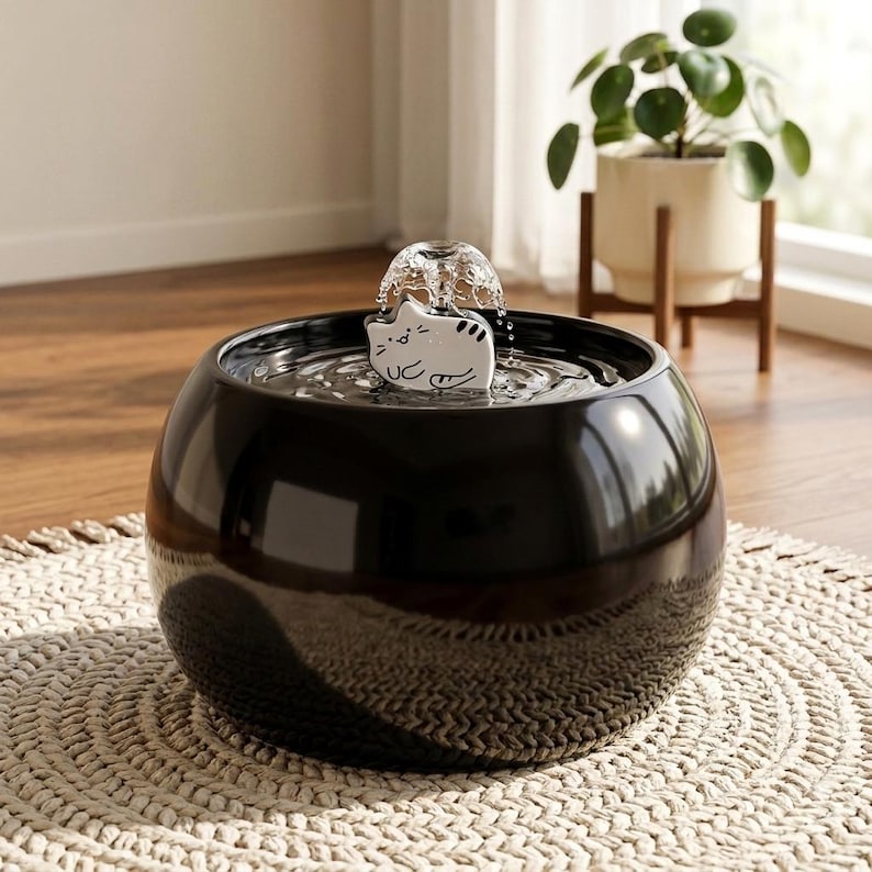 May include: A round, glossy black pet water fountain featuring a white ceramic cat-shaped attachment in the centre. The fountain creates a small, bubbling water dome effect at the top, providing a continuous flow of water for pets to drink from.