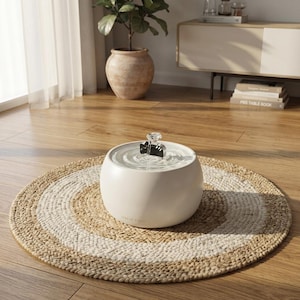 May include: A round, off-white pet water fountain with a smooth, matte finish. The fountain features a central black cat-shaped attachment where water flows, creating ripples on the surface. The brand name Serene Paws is printed on the front.