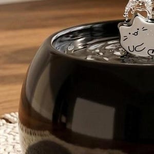May include: A glossy black ceramic pet water fountain with a rounded bowl shape. A white ceramic attachment shaped like a stylised cat face with simple black line features sits at the top, where water flows out into the basin below.