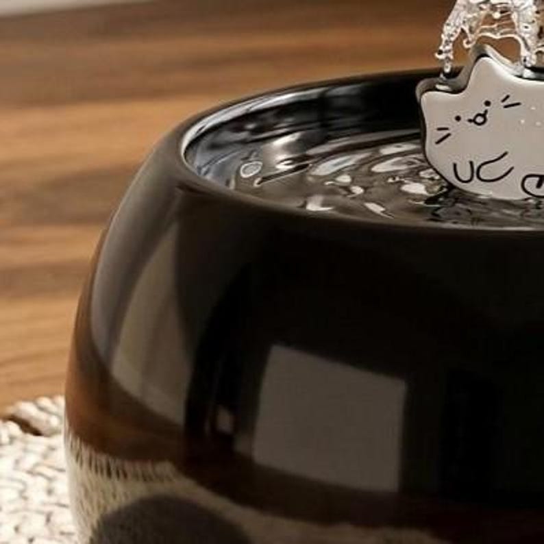 May include: A glossy black ceramic pet water fountain with a rounded bowl shape. A white ceramic attachment shaped like a stylised cat face with simple black line features sits at the top, where water flows out into the basin below.