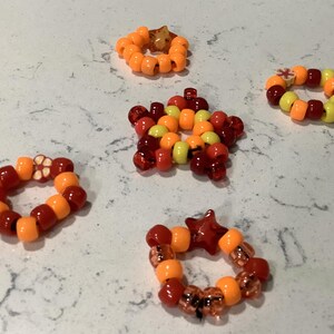 May include: A collection of five handmade rings crafted from colorful plastic pony beads in shades of orange, red, and yellow. The rings feature various bead shapes, including round, star-shaped, and flower-shaped accents, arranged in circular and star patterns.