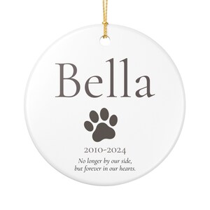 Può includere: Ornamento a disco in ceramica bianca con cordino dorato per appenderlo. Presenta il nome "Bella" in grigio scuro, un'impronta di zampa e gli anni 2010-2024. Il testo sottostante recita: "Non più al nostro fianco, ma per sempre nei nostri cuori."
