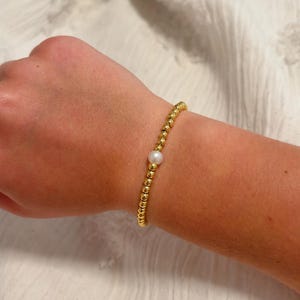 Puede incluir: Pulsera de cuentas doradas con un solo acento de perla blanca. La pulsera está hecha de pequeñas cuentas redondas doradas y una perla central redonda. La pulsera se usa en una muñeca sobre un fondo de tela blanca.