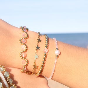 May include: Several beaded bracelets in different designs. One bracelet has gold beads and pink crystals in a wavy pattern. Another features gold star charms. A third is strung with small pink beads and a single pearl.