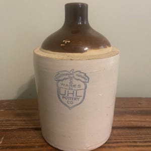Könnte beinhalten: Ein Vintage-Steinzeugkrug mit braunem Deckel und cremefarbenem Körper. Der Krug hat ein blaues Emblem mit dem Text "UHL Pottery Co. Huntingburg, Indiana". Der Krug wird wahrscheinlich zur Aufbewahrung von Flüssigkeiten verwendet.