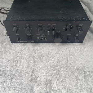 Könnte beinhalten: Schwarzer Audioverstärker mit mehreren Knöpfen und Reglern. Die Frontplatte verfügt über Bedienelemente für Bass, Höhen, Balance und Funktion. Die Oberseite des Geräts hat Lüftungsschlitze. Der Verstärker steht auf einer weißen, flauschigen Oberfläche.