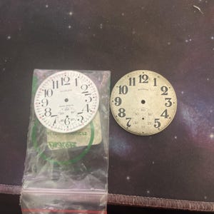 Może przedstawiać: Dwie zabytkowe tarcze zegarowe. Jedna jest biała z czarnymi cyframi i napisem "ILLINOIS" i "BUNS-SPECIAL". Druga jest w kolorze złamanej bieli z czarnymi cyframi i napisem "ILLINOIS". Oba mają małe tarcze dla sekund.