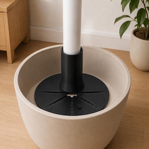 May include: A beige, bowl-shaped planter containing a black, circular, multi-slotted insert base with a central cylindrical sleeve. A white, vertical pole is inserted into the black sleeve, extending upward from the center of the planter.