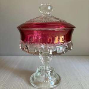 May include: A clear and cranberry red glass candy dish with a lid. The bowl features a raised hobnail pattern around the base and a smooth, deep red band around the upper rim. It stands on a pedestal base with a matching hobnail design.
