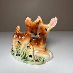 May include: A ceramic figurine featuring a standing fawn and a reclining deer. The animals are brown with white spots, black eyes, and pink-tinted ears. They are positioned on a base moulded to resemble green grass with a glossy, smooth finish.