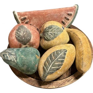 May include: A round wooden bowl containing a collection of decorative fruit sculptures, including a green pear, a red apple, a yellow mango, a yellow banana, and a large watermelon slice, all featuring carved leaf details and matte finishes.