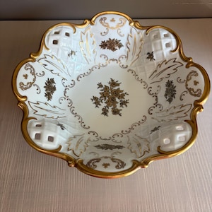 May include: A white porcelain bowl with a scalloped edge and gold trim. The bowl features a central floral motif in gold, surrounded by gold leaf patterns and lattice-style cutouts along the rim. The design includes intricate gold filigree details.