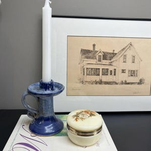 May include: A blue ceramic candle holder with a white taper candle, a small round cream-coloured lidded trinket box with a floral design, and a book titled Women's Encyclopedia of Health & Emotional Healing, positioned before a framed house sketch.