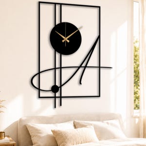 Puede incluir: Reloj de pared de metal negro con manecillas doradas. El reloj está montado en una pared blanca y presenta un diseño abstracto con un marco rectangular, una esfera circular y una letra "A" estilizada.