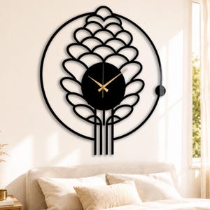 May include: Black metal wall clock with a unique design. The clock features a circular face with gold-colored hands, surrounded by a stylized floral or plant-like silhouette. The clock is mounted on a white wall above a bed.