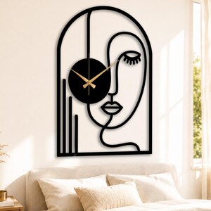 Könnte beinhalten: Schwarze Metall-Wanduhr in einem abstrakten Gesichtsdesign. Die Uhr hat ein schwarzes kreisförmiges Zifferblatt mit goldfarbenen Zeigern. Das Gesicht ist in eine größere Umrisslinie eines Gesichts mit gewölbtem Oberteil integriert.