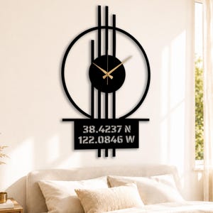 Op de afbeelding: Zwarte metalen wandklok met een cirkelvormig ontwerp, verticale staven en een rechthoekige basis. De wijzerplaat is zwart met goudkleurige wijzers. De basis toont de coördinaten 38.4237 N 122.0846 W. Decoratieve wandkunst.