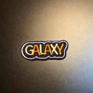 May include: An embroidered patch featuring the word GALAXY in alternating yellow and orange thread. The text is set against a black background with a thick, wavy lavender border that follows the contour of the letters in a rounded shape.