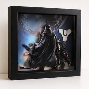 May include: A square black shadow box frame containing a multi-layered 3D paper art display. The artwork features three armoured figures in dark cloaks standing before a large blue planet, with the white Destiny game logo visible on the right side.