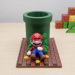 以下が含まれることがあります： 緑色の円筒形パイプ型ペン立て。赤いレンガ模様の長方形の台座の上に、Mのロゴ入り赤い帽子、赤いシャツ、青いオーバーオールを着たマリオのフィギュアが座っており、周囲には金色のコインと緑色の茂みが配置されています。
