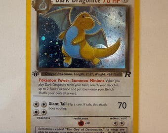 Può includere: Una carta collezionabile olografica Dark Dragonite con bordo giallo. La carta presenta una creatura gialla e blu, 70 HP, e il testo 'Pokémon Power: Summon Minions' e 'Giant Tail'. È una carta di prima edizione con un simbolo R nero.