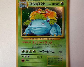 Può includere: Una carta collezionabile giapponese Venusaur con un'illustrazione olografica della creatura blu con un grande fiore arancione sul dorso. La carta mostra il nome Fushigibana, 100 HP e varie statistiche di gioco con simboli e testo.