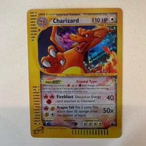 Könnte beinhalten: Eine holografische Charizard-Sammelkarte mit gelbem Rand, die den Charakter in einer dynamischen Pose zeigt. Die Karte weist 110 KP, die Fähigkeit Crystal Type sowie die Angriffe Fireblast und Dragon Tail auf, mit Copyright-Text von 2003.