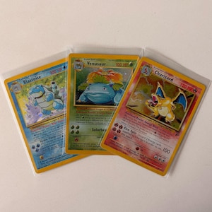 May include: Three collectible trading cards in protective plastic sleeves, featuring the Pokémon characters Blastoise, Venusaur, and Charizard. Each card displays the character illustration, name, HP value, and detailed game text in English.
