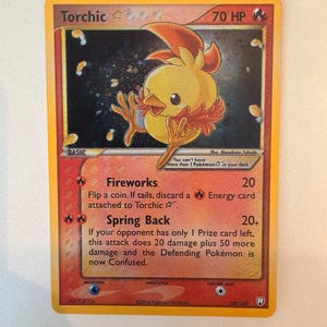 Puede incluir: Una carta coleccionable de Torchic que muestra una criatura amarilla similar a un pollito con detalles naranjas sobre un fondo oscuro y estrellado. La carta presenta 70 HP, los ataques Fireworks y Spring Back, y texto sobre mecánicas.