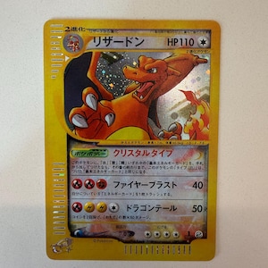 Puede incluir: Una carta coleccionable japonesa de Charizard con una ilustración holográfica de la criatura naranja. La carta muestra el nombre リザードン, 110 HP y dos ataques, Fire Blast y Dragon Tail, con símbolos de energía de fuego y rayo.