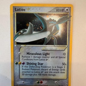 Puede incluir: Una carta coleccionable Pokémon Latios Star con 80 HP. La carta presenta una ilustración holográfica de la criatura Latios azul y blanca sobre un fondo oscuro y estrellado. Incluye los ataques Miraculous Light y Shining Star.
