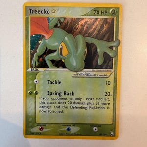 Puede incluir: Una carta coleccionable Pokémon Treecko con 70 HP, que muestra una ilustración de una criatura verde similar a un lagarto. La carta presenta los ataques Tackle y Spring Back, con texto sobre reglas, fecha de copyright 2004 y el número 109/109.