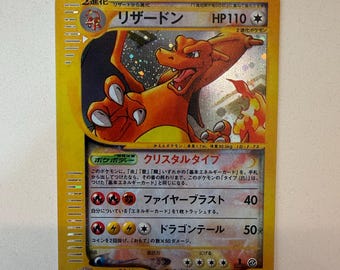 以下が含まれることがあります： オレンジ色のドラゴン風のキャラクターが描かれたホログラフィック仕様の日本語版リザードン・トレーディングカード。カードには「リザードン」、HP110、そして炎と雷のエネルギーシンボルを伴う「ファイヤーブラスト」と「ドラゴンテール」という2つの技が記載されています。