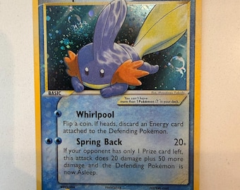 Può includere: Una carta collezionabile Pokémon Mudkip con 70 HP, caratterizzata da un'illustrazione olografica della creatura blu. La carta mostra gli attacchi Whirlpool e Spring Back, insieme al testo di gioco, un bordo giallo e uno sfondo blu.