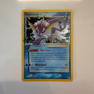 May include: A Vaporeon trading card with 70 HP, featuring a purple creature illustration, a blue background, and the text Blue Ray and Whirlpool. The card has a yellow border and holographic details showing water bubbles around the character.