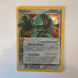May include: A Registeel trading card featuring a metallic green creature with a dark face and red dots. The card displays 90 HP, the attacks Barrier Attack and Final Laser, and includes text detailing game mechanics, set information, and copyright.