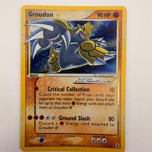 Puede incluir: Una carta coleccionable Pokémon Groudon Gold Star con 90 HP. La carta presenta una ilustración de un Groudon oscuro sobre un fondo estrellado, con los ataques Critical Collection y Ground Slash listados bajo la ilustración sobre un fondo naranja.