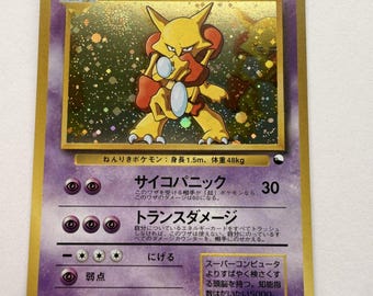 Può includere: Una carta collezionabile olografica di Alakazam con bordo viola e cornice dorata. La carta raffigura il personaggio giallo che tiene due cucchiai d'argento, con testo giapponese, HP90 e simboli di energia psichica sul fronte della carta.