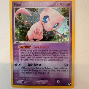 May include: A holographic Mew trading card with 70 HP, featuring a pink Mew character illustration. The card displays the Poke-Power Type Change and the Link Blast attack with 50 damage. It includes text details, a psychic energy symbol, and a yellow border.