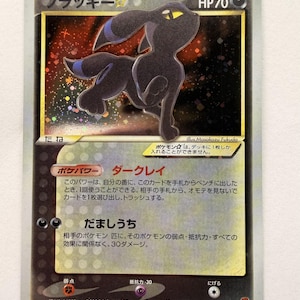 Puede incluir: Una carta coleccionable Pokémon Umbreon Gold Star japonesa de 2005 que muestra una criatura negra con ojos amarillos y anillos azules sobre un fondo holográfico oscuro. La carta muestra 70 HP, texto en japonés y el nombre Masakazu Fukuda.