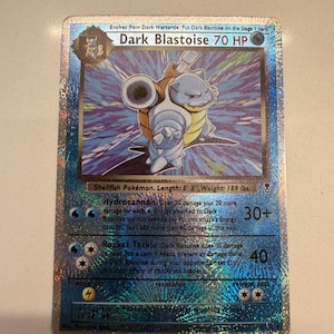 Pode incluir: Uma carta colecionável holográfica Dark Blastoise com 70 HP. A carta mostra o personagem com uma carapaça azul e bege, incluindo os ataques Hydrocannon e Rocket Tackle. O texto inclui Shellfish Pokémon, 5' 3", 189 lbs.