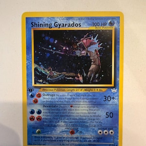 May include: A Shining Gyarados trading card with a yellow border and blue background. The illustration shows a red sea serpent creature emerging from dark water near a small boat. Text includes 100 HP, Outrage, Devastate, and 1st Edition symbol.
