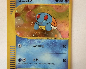 Può includere: Una carta collezionabile Pokémon Squirtle giapponese con bordo giallo e un'illustrazione olografica del personaggio tartaruga blu. La carta mostra HP50, il testo "ゼニガメ", "ぶつかる" e "あわ", insieme a "身長0.5m、体重9kg" e il logo McDonald's nell'angolo in basso a destra.