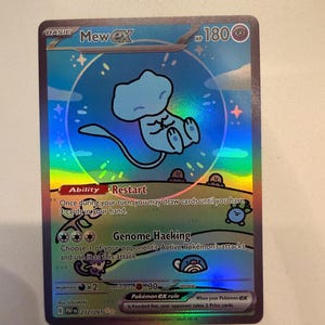 Op de afbeelding: Een Mew ex ruilkaart met een holografische afwerking. De kaart toont het personage Mew zwevend in een blauwe bubbel boven een graslandschap met kleine wezens. De tekst bevat Mew ex, 180 HP, Ability Restart en Genome Hacking.